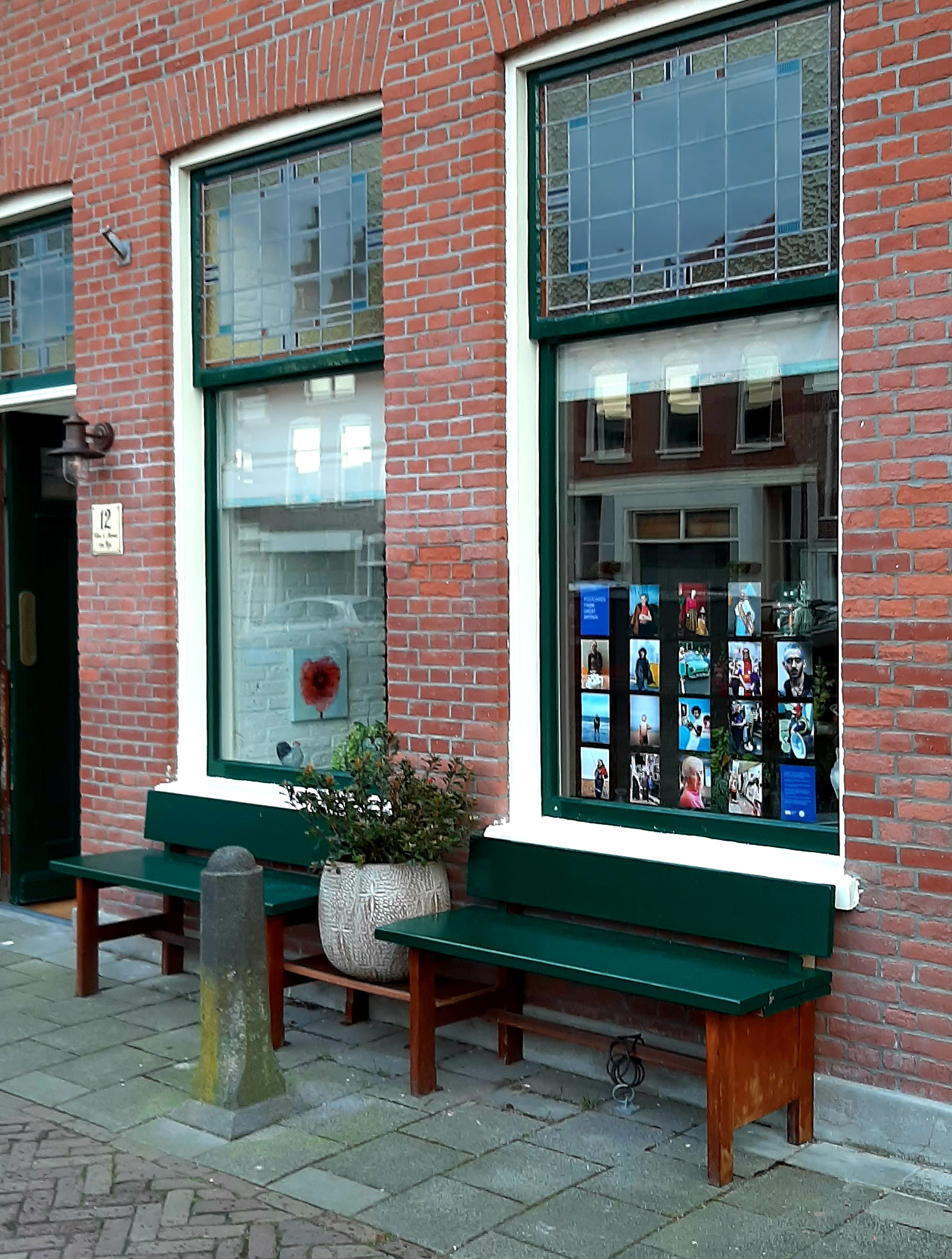 Photograph of two windows, the one to the right displaying postcard-sized photographs, with brick wall either side, a green wooden bench in front of both windows and a potted plant and a concrete pillar in between.