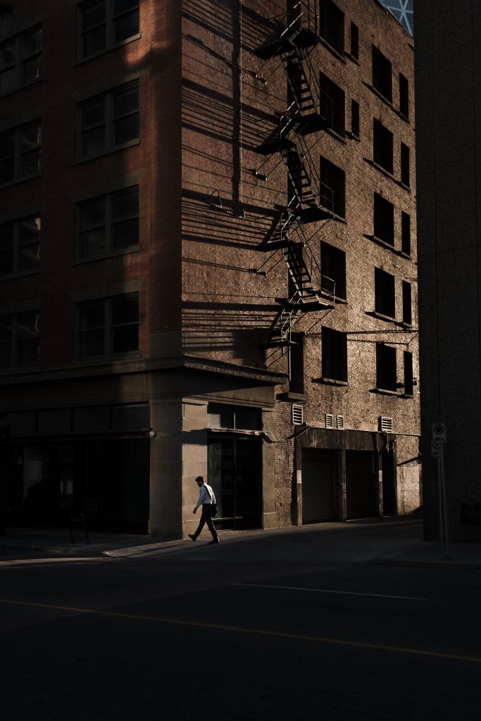Person walks into a building’s shadow while the brick side and fire-escape are lit by the sun.