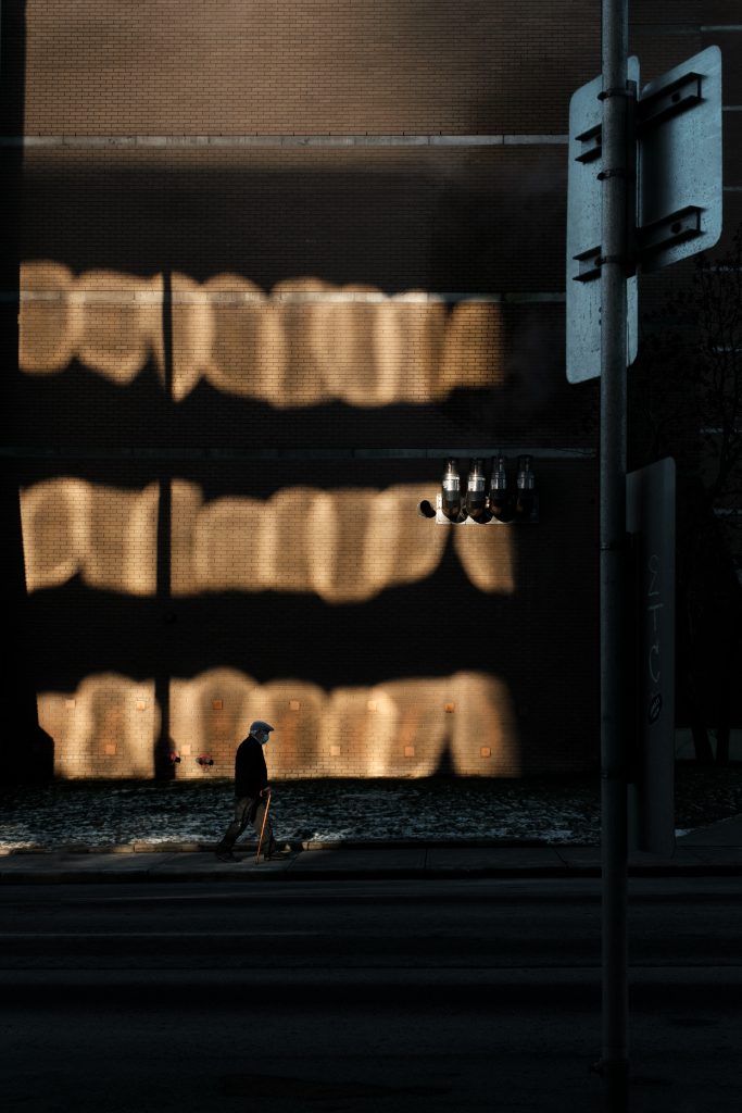 An elderly person walks with a cane through reflected light on the street.