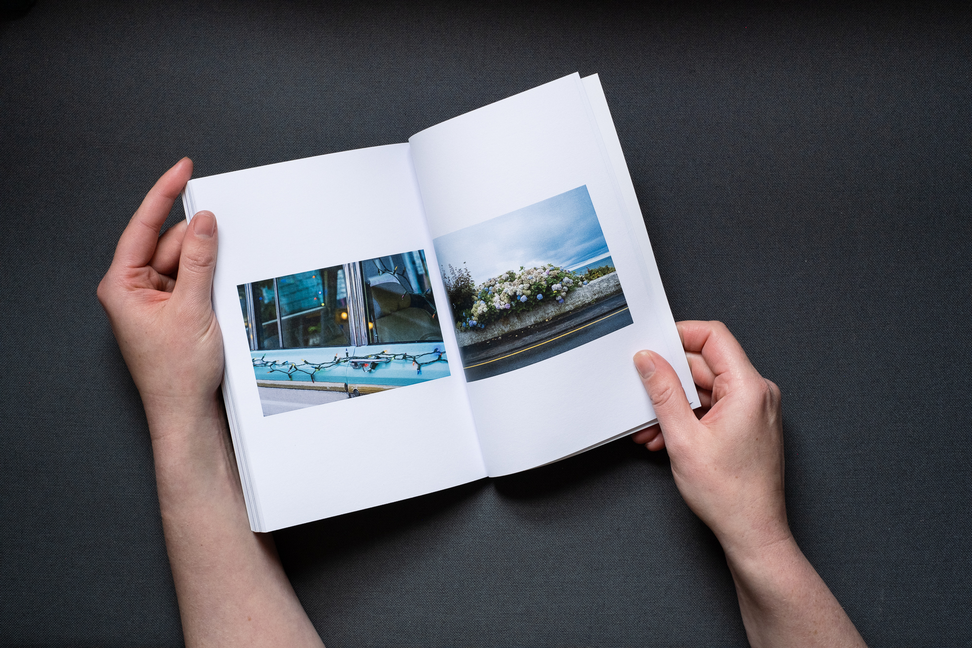 Photograph of the ROAD TRIP printed publication being held open by hands, on a dark grey background. The left page shows a photograph looking through turquoise grand wagoneer windows with fake wood paneling and small christmas lights and reflections. The right page shows a photograph of a hydrangea bush at the roadside, with the flowers changing colour, including blue, pink, lilac and brown.
