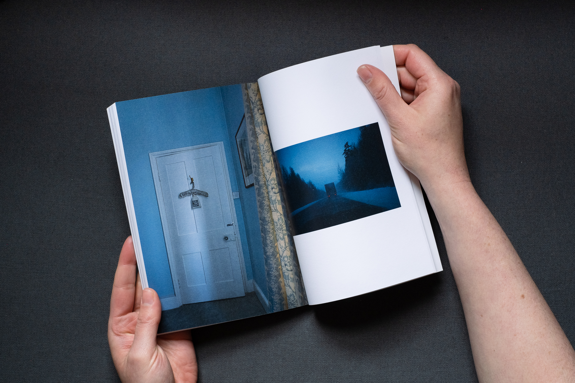 Photograph of the ROAD TRIP printed publication being held open by hands, on a dark grey background. The left page shows a photograph of a coat hanger on back of sunlit bedroom door framed by blue curtains and blue walls. The right page shows yhe back of a truck seen in the distance in the low light of a snowy forest road.