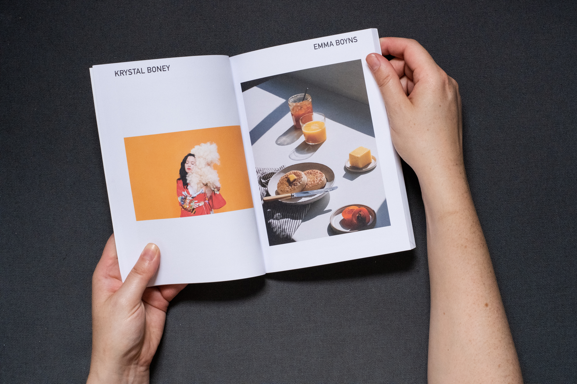 Photograph of hands holding a book against a dark grey fabric background. A double page spread is shown, on the left page text says 'KRYSTAL BONEY' at the top and there is a photograph below of a woman in a red kimono hugging a blonde curly wig against an orange background. On the right page text says 'EMMA BOYNS' with a photograph below of a plate of crumpets resting on a striped tea towel with melting butter on top, on a table by a window. A jar of marmalade and juice on the left and a halved apricot and square of butter on the right. Harsh sunlight creates strong shadows.