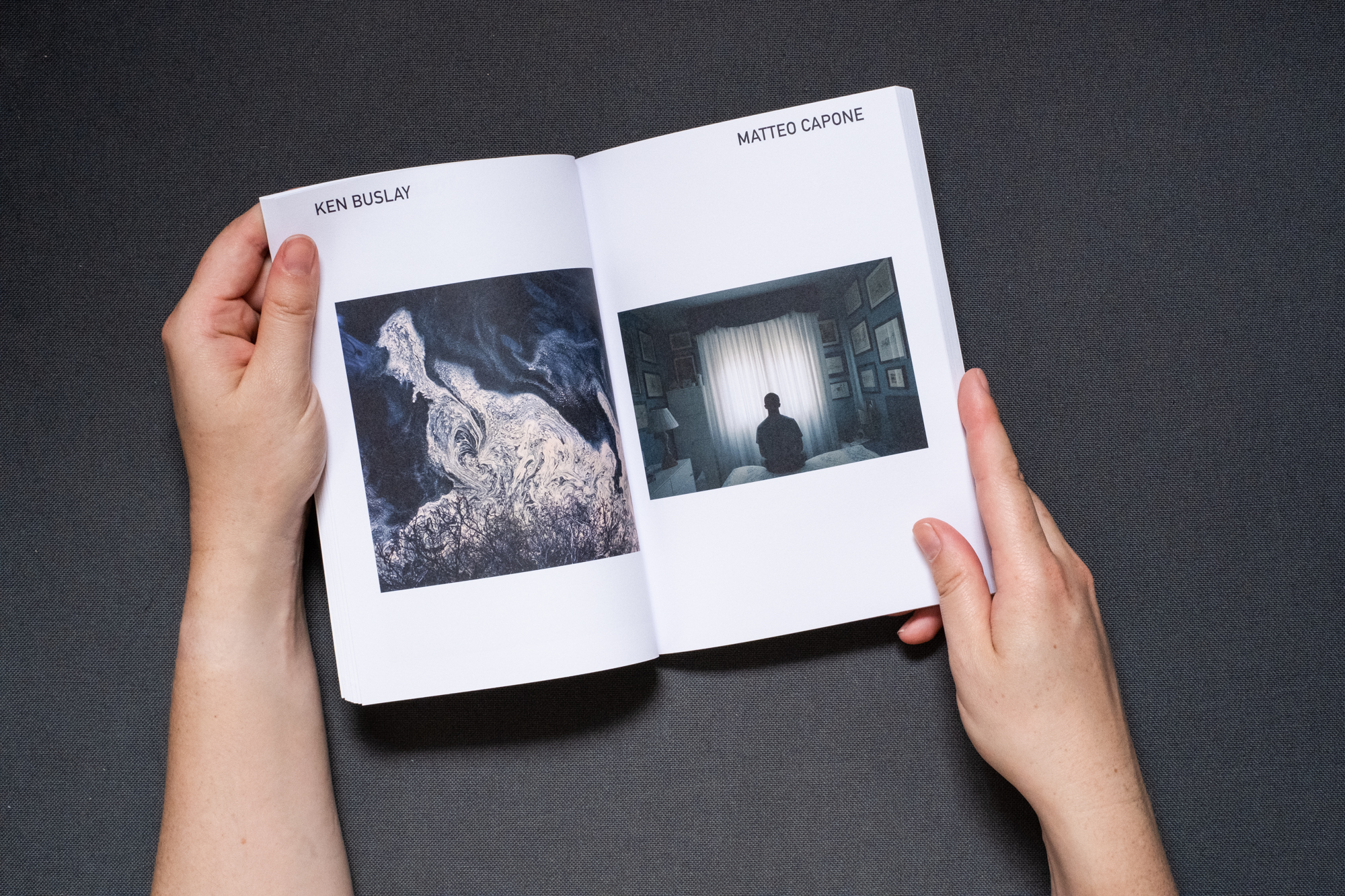 Photograph of hands holding a book against a dark grey fabric background. A double page spread is shown, on the left page text says 'KEN BUSLAY' at the top and there is a photograph below of Pollen on water forming intertwining structures. On the right page text says 'MATTEO CAPONE' with a photograph below of self-portrait in a room during the second Italian lockdown.