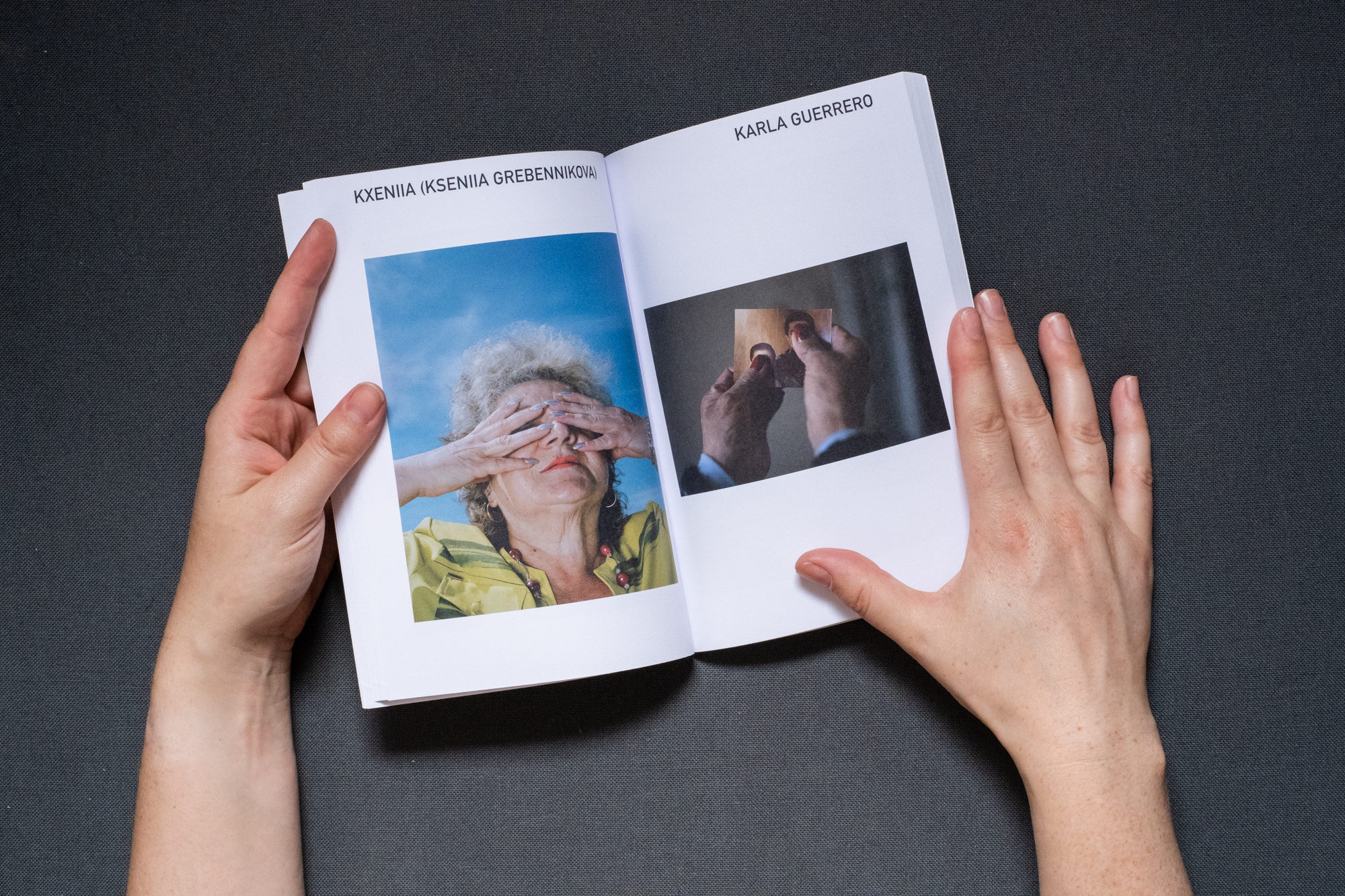 Photograph of hands holding a book against a dark grey fabric background. A double page spread is shown, on the left page text says 'KXENIIA (KSENIIA GREBENNIKOVA)' and there is a photograph below of the photographer's grandmother covering her face with her arms with a blue sky on the background. On the right page text says 'KARLA GUERRERO' and there is a photograph below of the hands of an old woman holding a photography in which she covers the people portrayed in the photograph.