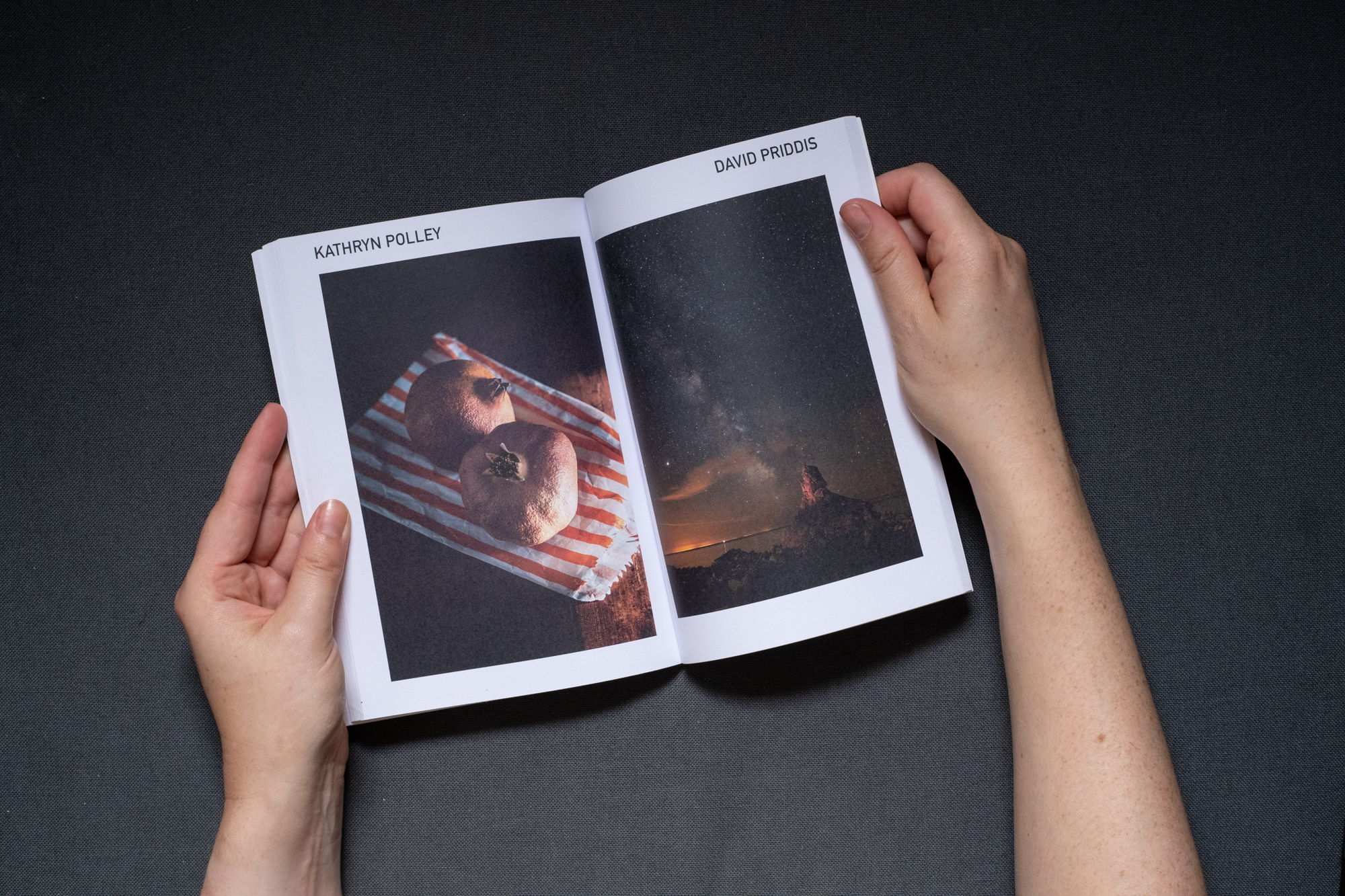 Photograph of hands holding a book against a dark grey fabric background. A double page spread is shown, on the left page text says 'KATHRYN POLLEY' and there is a photograph below of two dried out pomegranates sitting on top of an orange and white striped paper bag. On the right page text says 'DAVID PRIDDIS' and there is a photograph below of a star filled night sky with the milkyway running diagonally across and ending at a rock stack in the sea.