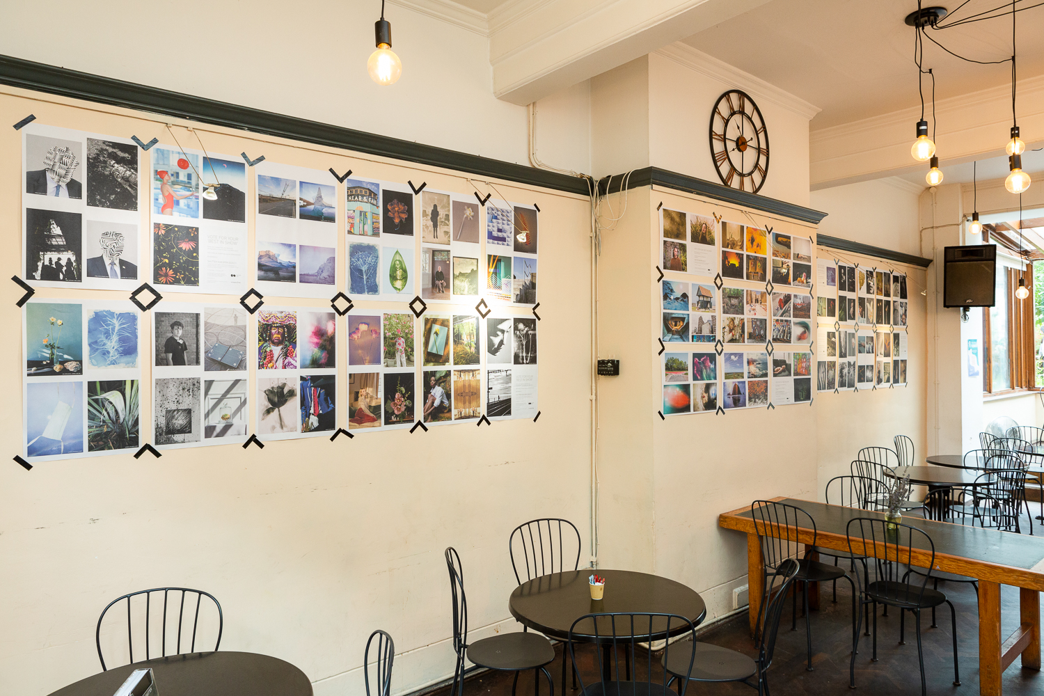Photograph in the Gallery Café at St. Margaret's House, showing an exhibition of photographs printed on newsprint, tables and chairs in the foreground