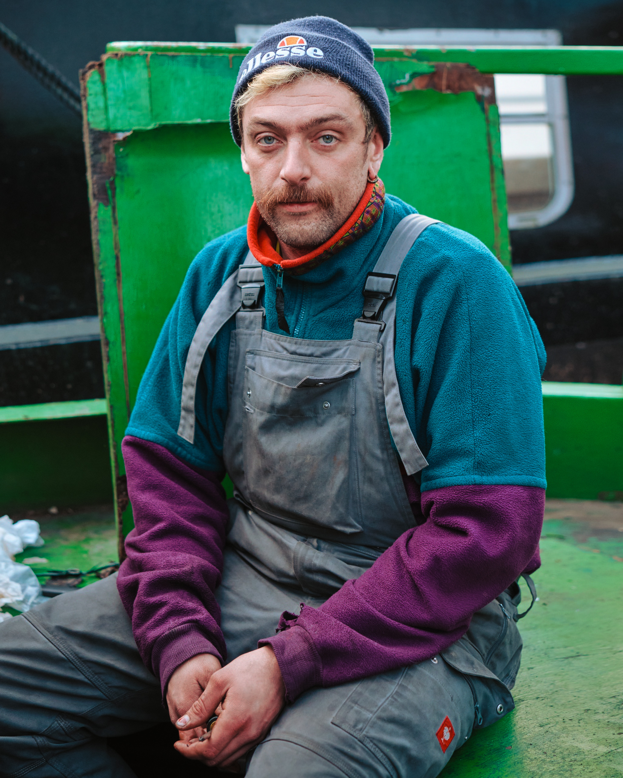 portrait of a man in grey dungarees sat on the back of a green narrowboat.