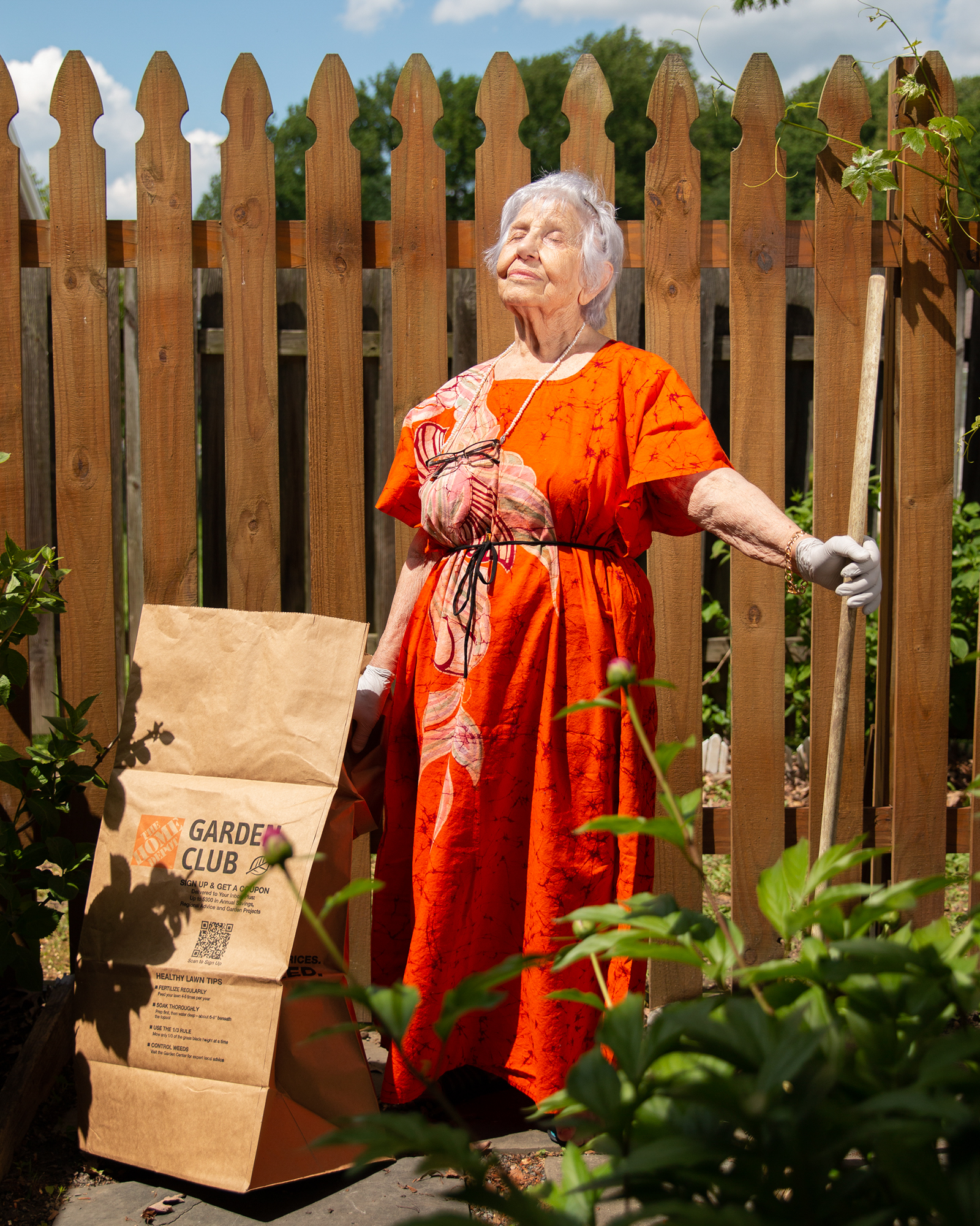 A woman in her 80s stands outside on a sunny day. She is wearing an orange dress and is holding a rake in her left hand. A garden clippings bag is on the ground to her right. Her face is tilted upward towards the sun and her eyes are closed.