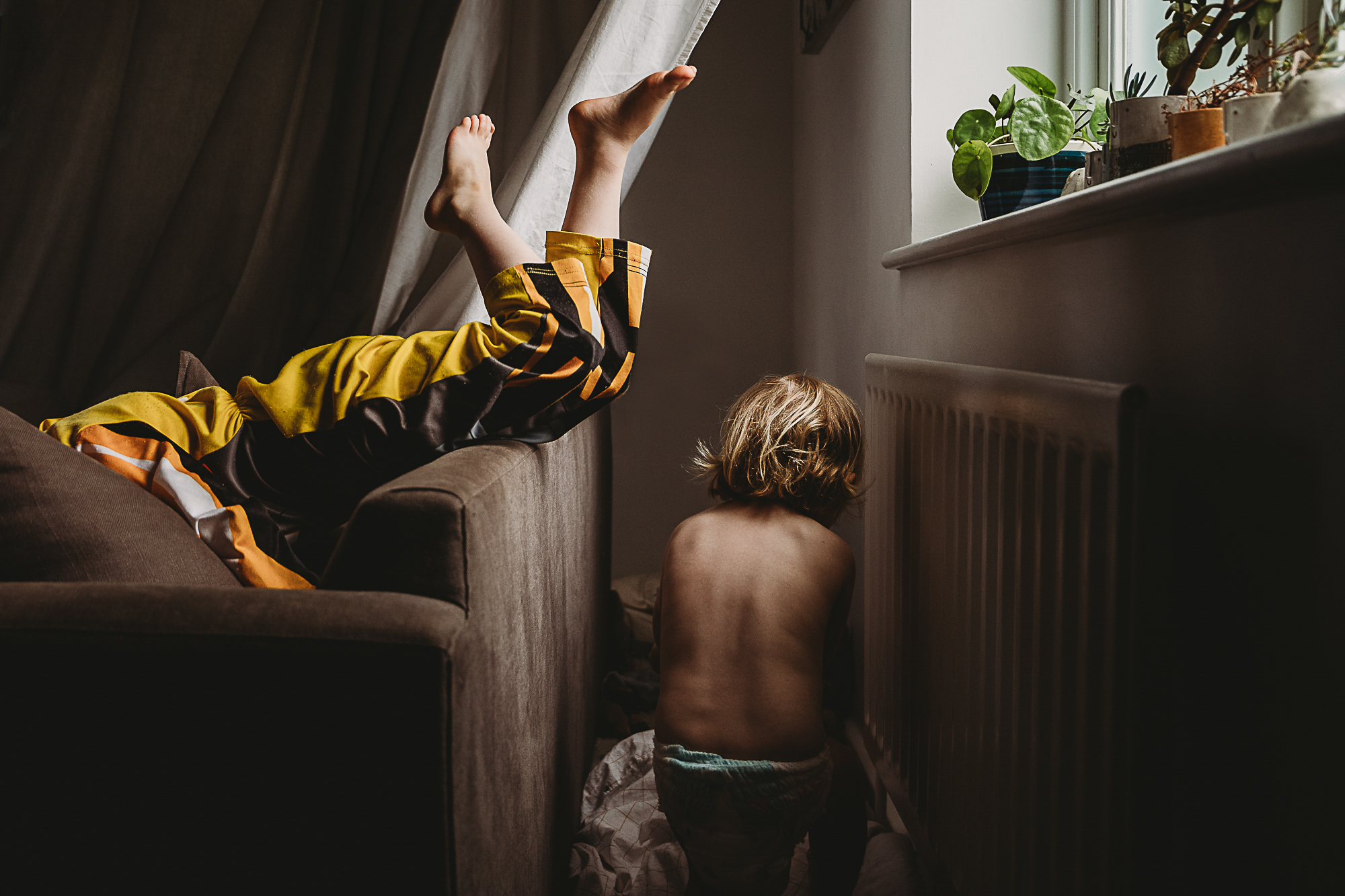 Boy wearing yellow superhero costume leaps over a sofa, with just his legs visible, while his younger sister runs to hide behind a sofa.