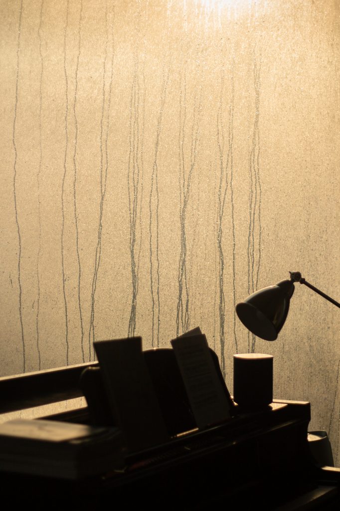 A grand piano with music scores and a lamp is silhouetted against a large window which is running with condensation back lit by sunlight.